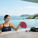 Woman holding a red wine LUWA Apex swim cap by a pool. Premium silicone cap for comfort and durability.